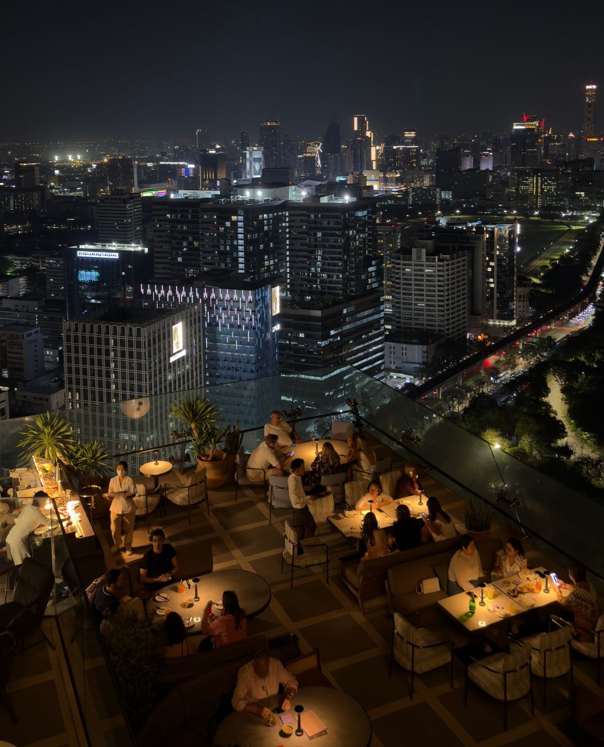 แขกกำลังรับประทานอาหารที่โต๊ะซึ่งมีแสงไฟส่องสว่างบนระเบียงร้านอาหารบนชั้นดาดฟ้าในกรุงเทพฯ ยามค่ำคืน โดยมีเส้นขอบฟ้าของเมืองที่สว่างไสวอยู่นอกระเบียงกระจก