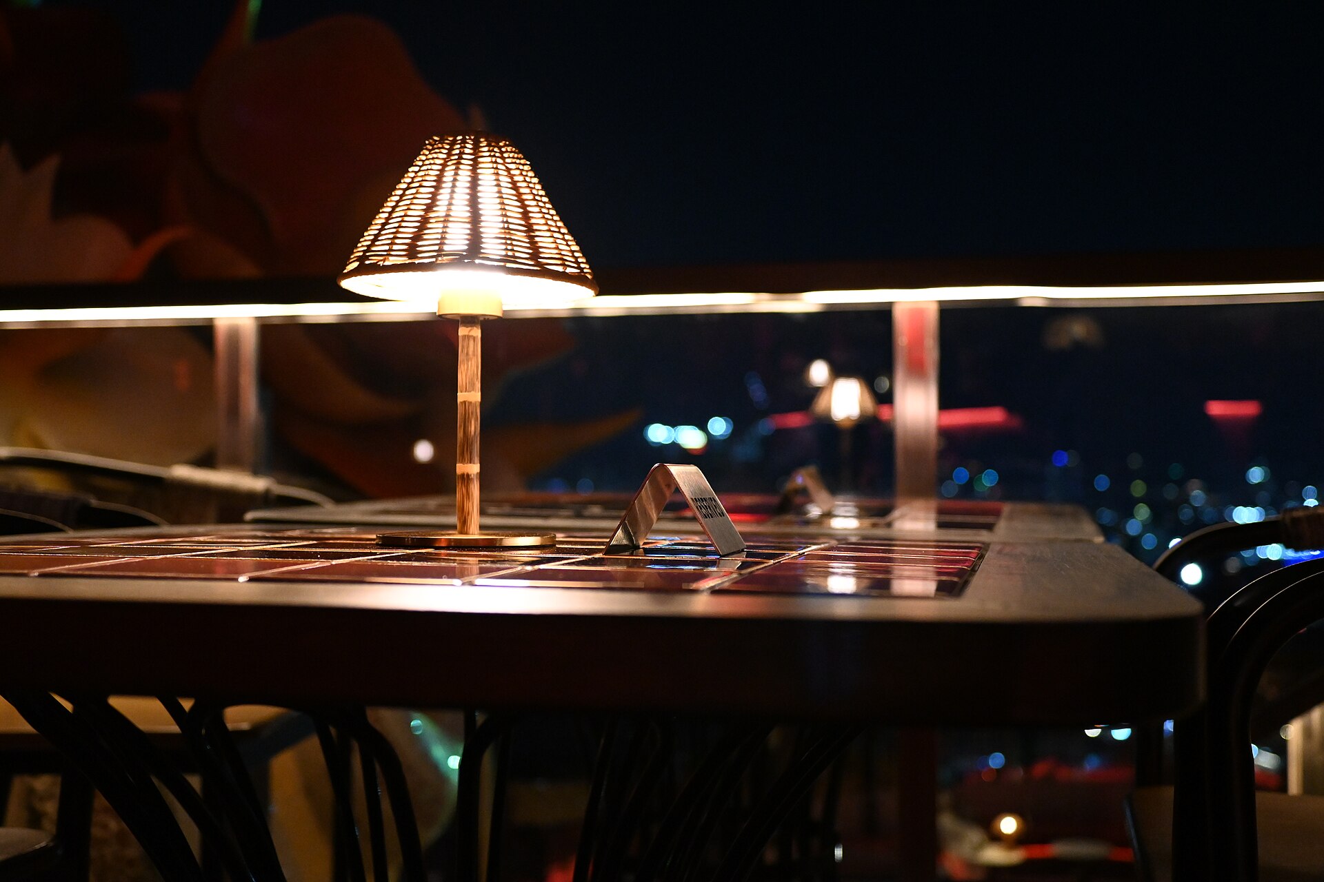 A lamp-lit tiled dining table on a rooftop terrace at night, chairs drawn up and Pattaya's city lights spread out below the glass balustrade